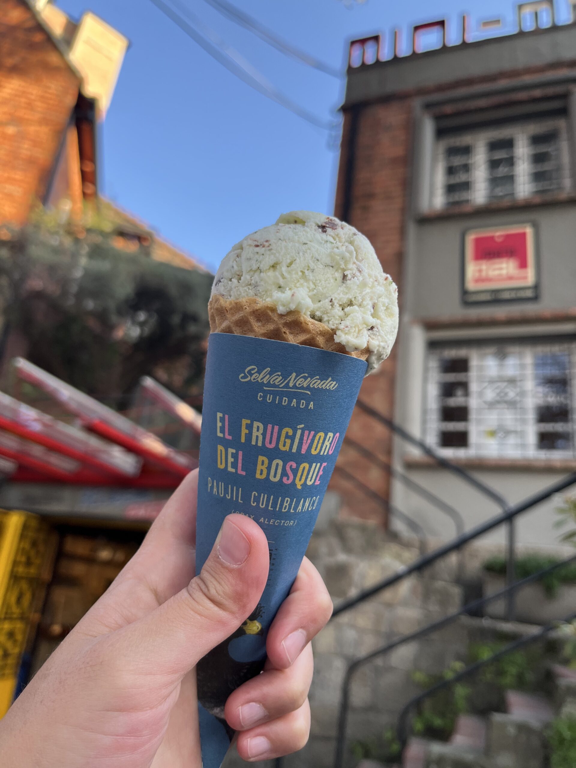 A hand holding an ice cream cone in front of a building. The cone has a blue wrapper with text in Spanish, including “El Frugívoro del Bosque.” It's one of those delightful things to do in Bogotá, making any tour of the city extra sweet.