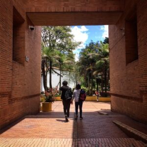 Two people, one with a black backpack and the other with a red backpack, walk through a brick archway towards a garden area filled with palm trees and plants under a bright blue sky—just one of the many delightful experiences offered on Bogotá city tours.