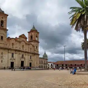 Historical stone church with twin towers in a large plaza. Surrounding the plaza are colonial-style buildings, a tall palm tree, and people exploring the area. This is a must-see on any Bogotá city tour. The sky is partly cloudy.