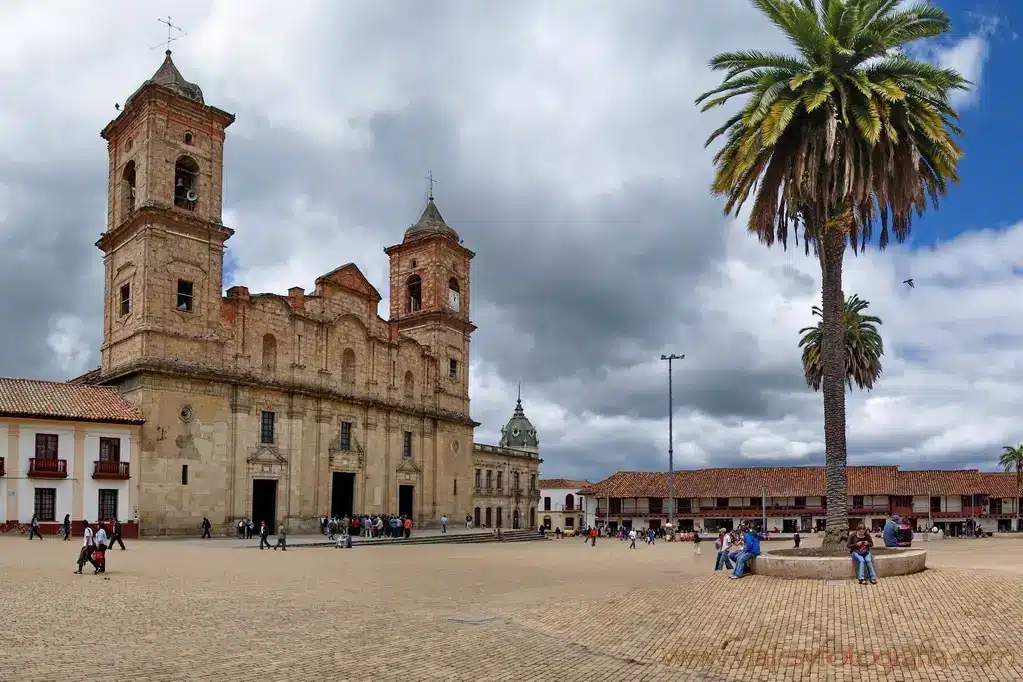 Historical stone church with twin towers in a large plaza. Surrounding the plaza are colonial-style buildings, a tall palm tree, and people exploring the area. This is a must-see on any Bogotá city tour. The sky is partly cloudy.