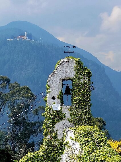 Old stone bell tower covered in ivy, with mountains and a distant building in the background. A hidden gem often highlighted in Bogotá city tours, it's a must-see for those exploring the things to do in Bogotá.
