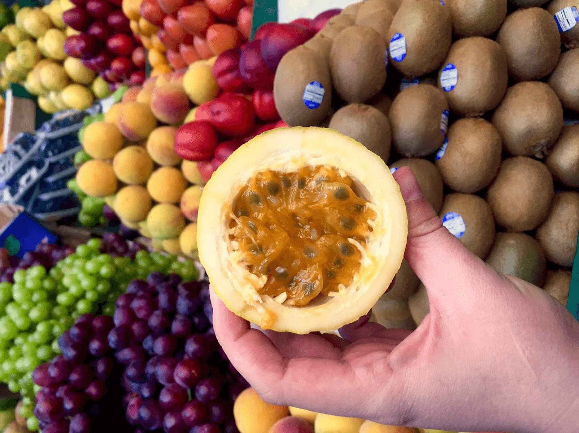A hand holds a halved passion fruit with seeds visible. In the background, there are various fruits including kiwis, grapes, and peaches displayed at a market—a perfect snapshot of the vibrant produce you can explore on Bogotá city tours.