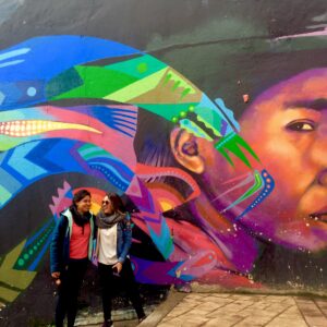 Two people stand in front of a vibrant mural featuring a close-up portrait with colorful, abstract patterns. They appear to be engaged in conversation and are smiling, enjoying one of the popular things to do in Bogotá during their city tour.