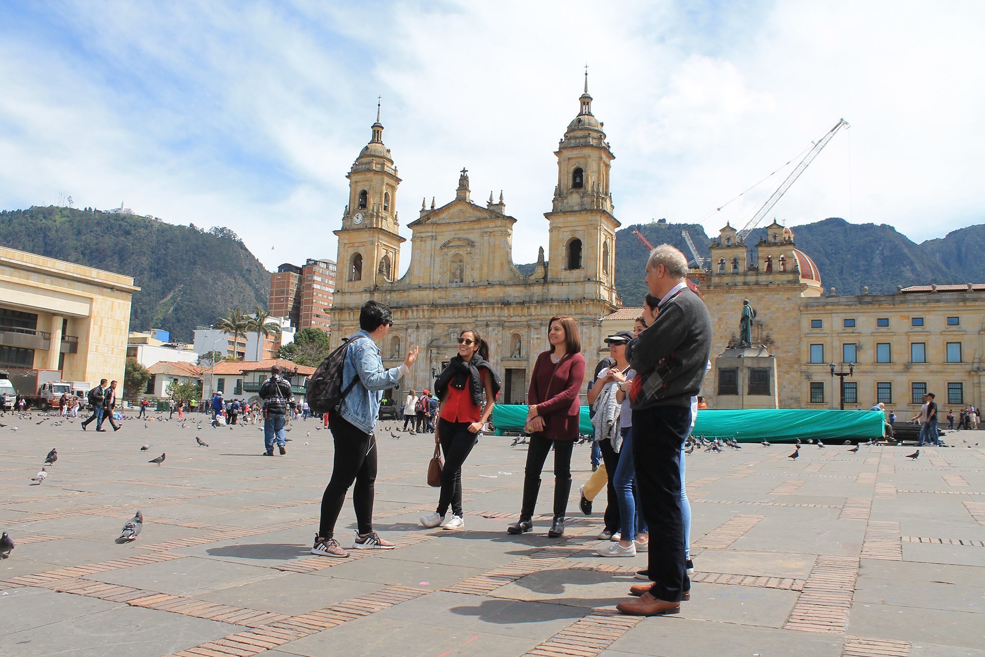 A group of people stands in a plaza in front of a historic building with two towers, surrounded by pigeons. A crane is visible in the background, and mountains can be seen in the distance—a perfect snapshot from one of many Bogotá city tours.