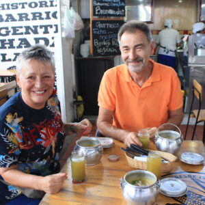 Two people seated at a table, smiling, with a handwritten menu behind them and two metal teapots and glasses of greenish liquid in front. Looks like they've just returned from an exciting Bogotá city tour. Indoor setting with another person cooking in the background.