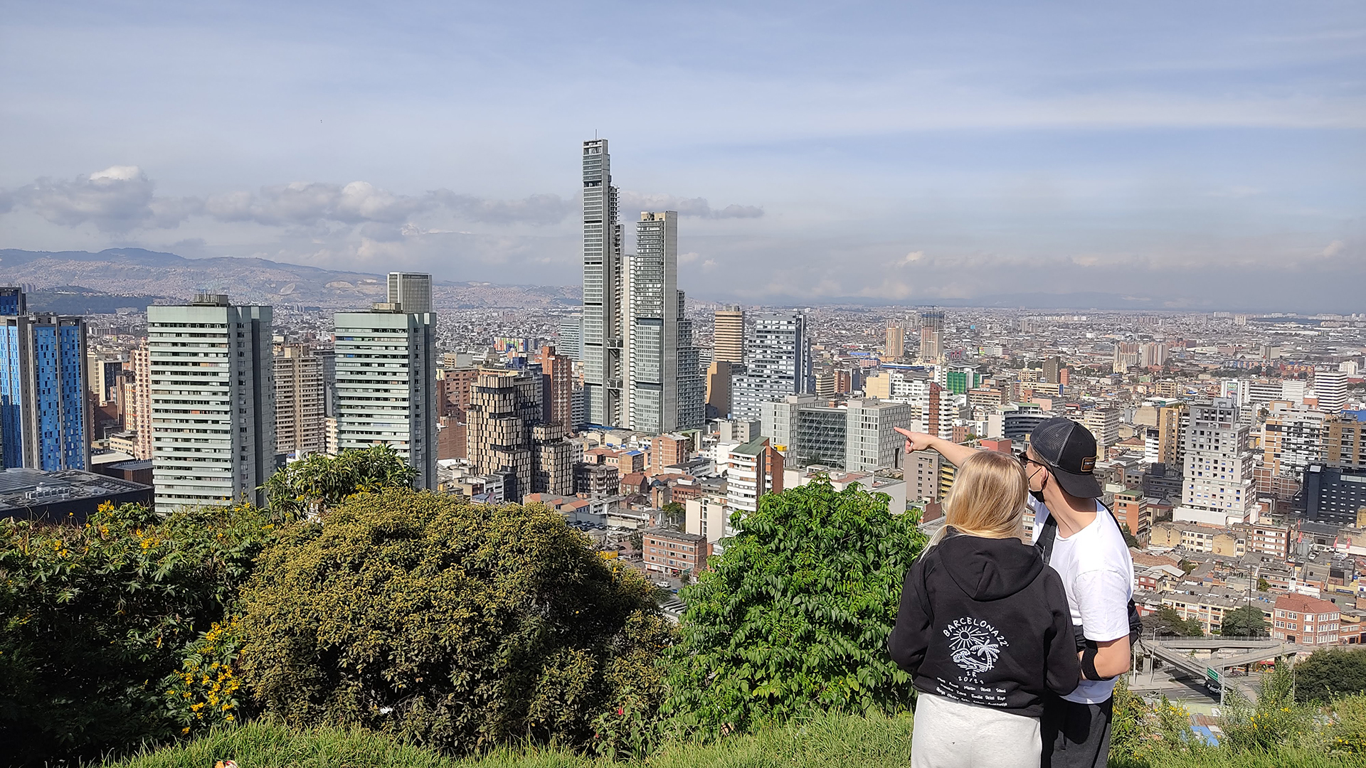A couple stands on a grassy hill overlooking Bogotá's cityscape filled with modern high-rise buildings. One person is pointing towards the skyline, suggesting one of the many tours in Bogotá as a must-do activity.
