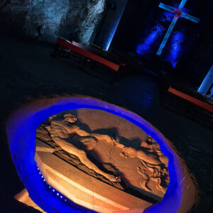 An illuminated circular relief sculpture depicting a classical scene is embedded in the ground of a dimly lit stone chamber, with a large, backlit cross visible in the background. This hidden gem is one of the unique things to do in Bogotá, making it a captivating stop on any Bogotá city tour.