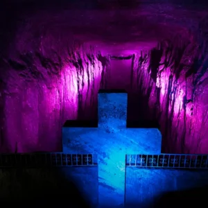 A large stone cross, illuminated by purple and blue lights, stands majestically in an underground cavern with rock walls. One of the captivating things to do in Bogotá includes exploring this mystical site on various Bogotá city tours.