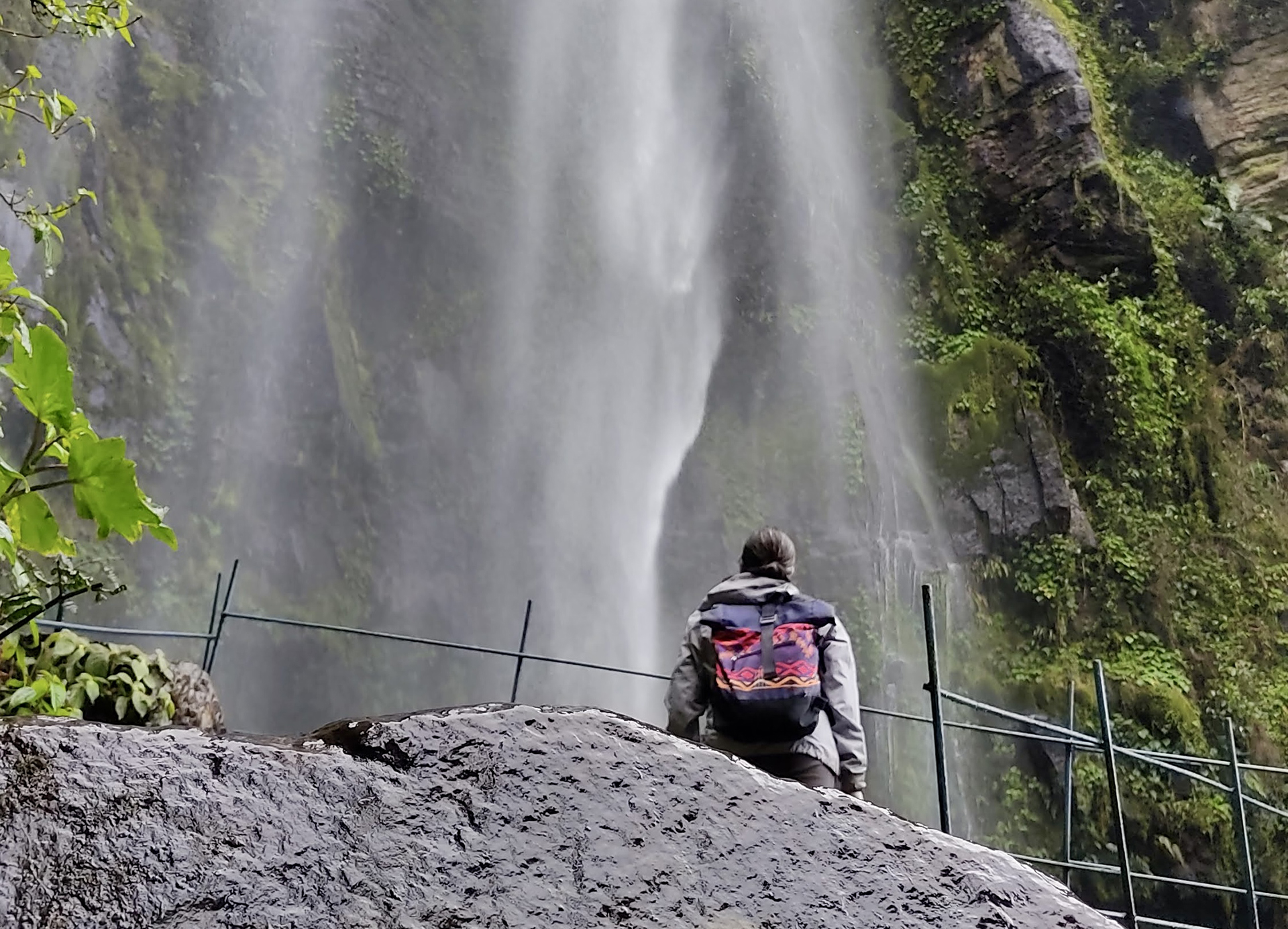 Individual with a colorful backpack stands on a rock near a tall waterfall, which cascades down a moss-covered cliff. A fence is visible next to them. This serene moment could easily be part of the many breathtaking sights offered by Bogotá city tours.