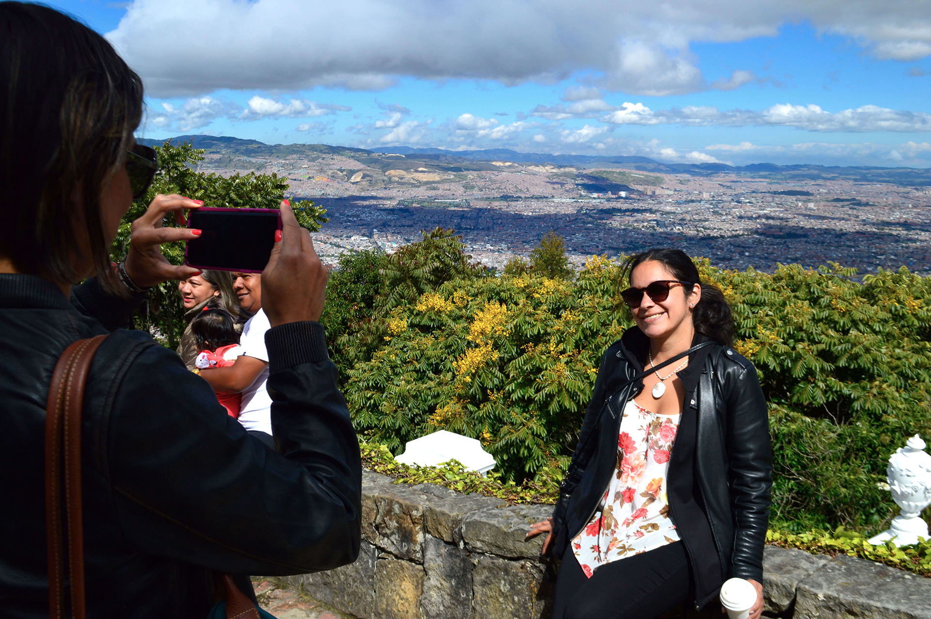 A person takes a photograph of a smiling woman in a floral top and leather jacket standing by a stone wall with a panoramic view of the city and mountains of Bogotá in the background, capturing one of the many highlights you can experience on Bogota city tours.
